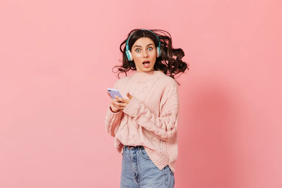Una mujer con auriculares mira su teléfono sobre un fondo rosa
